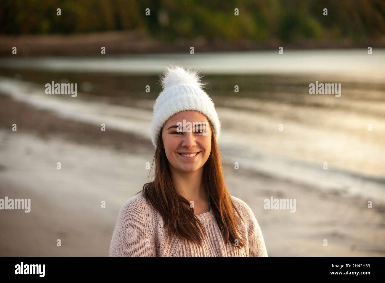 Eine Frau, die einen weißen Wollhut an einem Strand trägt und lächelt und zur Kamera schaut Stockfoto