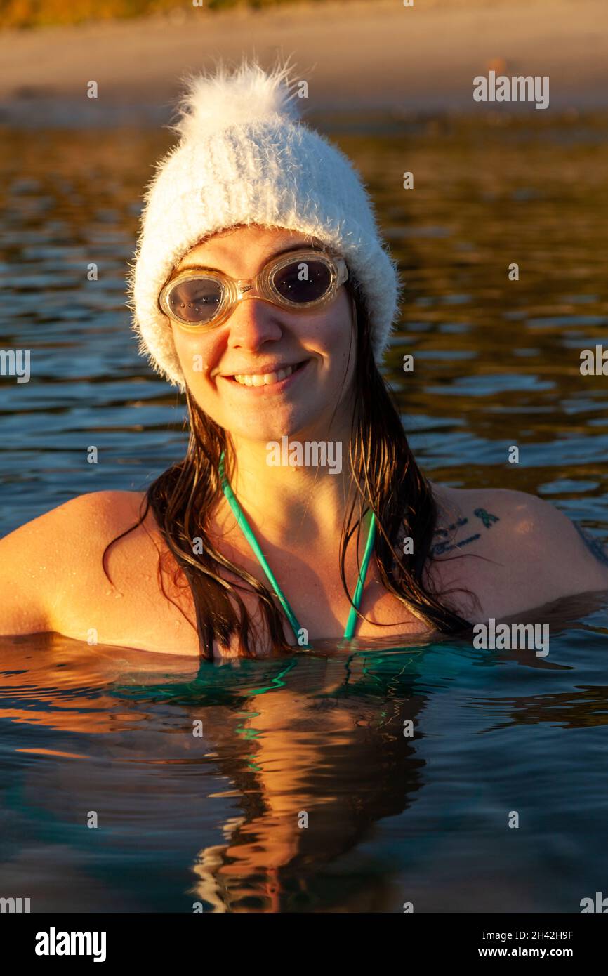 Eine junge schottische Frau, die im Oktober in Aberdour, Fife, Schottland, im Meer schwimmt und einen weißen Wollhut mit kaltem Wasser trägt Stockfoto