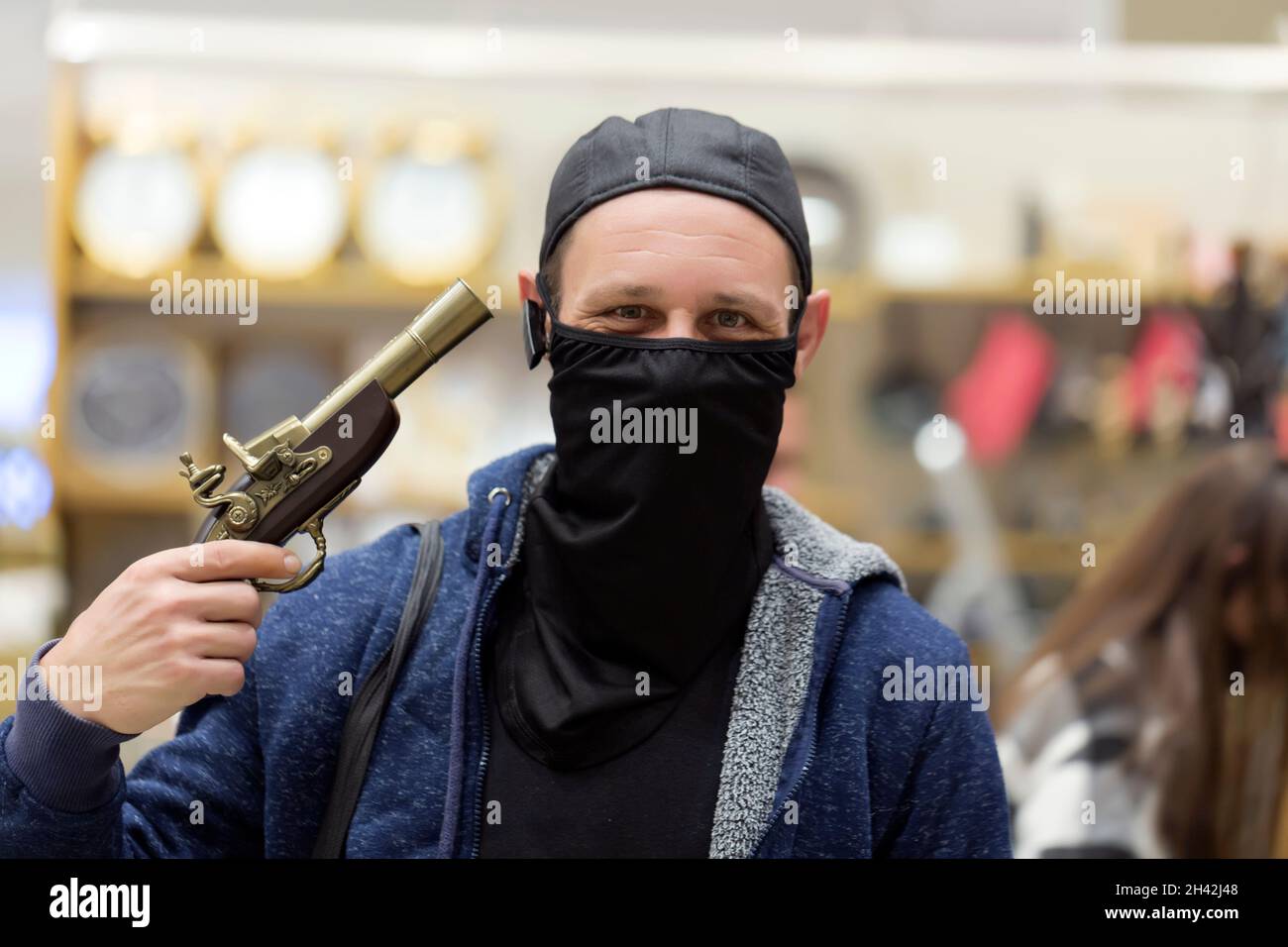 Kaukasischer Mann mit blauem Hoodie vor verschwommenem Hintergrund mit einer Waffe am Kopf. Mann mit schwarzer Gesichtsmaske. Banditen und Gangster. Stockfoto