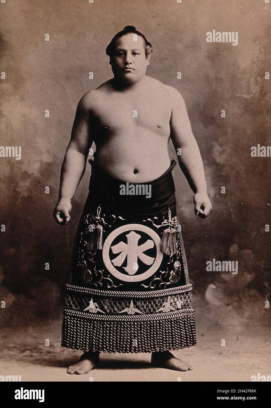 Ein junger Sumo-Wrestler, der in einem Fotostudio posiert, trägt eine reich verzierte Schürze vor einem gemalten Hintergrund. Foto, ca. 1900. Stockfoto
