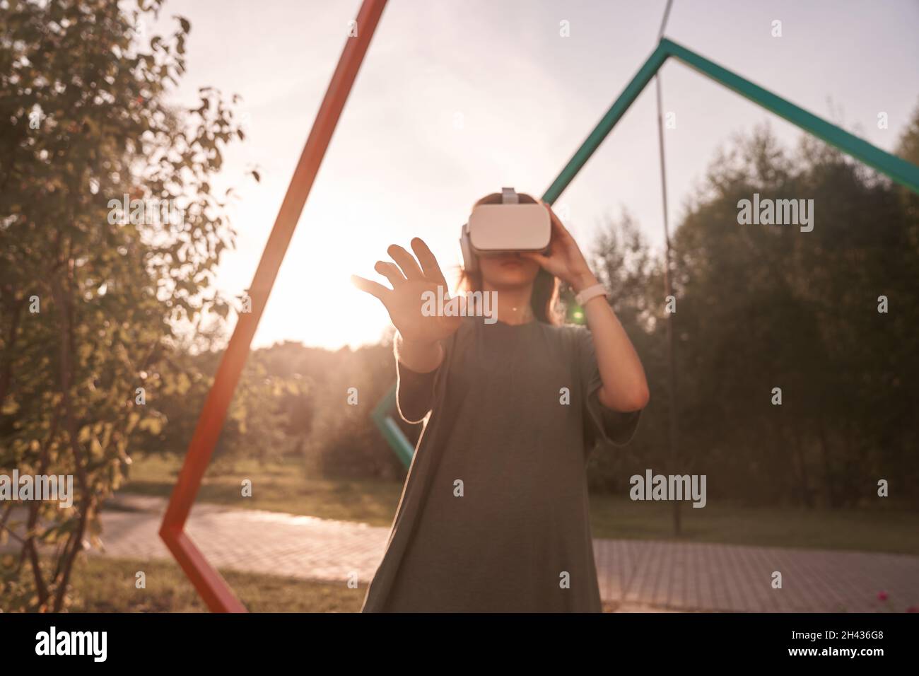 Nahaufnahme eines Mädchens, das mit den Händen eine Virtual-Reality-Brille auf dem Kopf hält und auf einem Hintergrund von grünen Bäumen steht. Er reicht eine Hand an die Kamera. Hochwertige Fotos Stockfoto