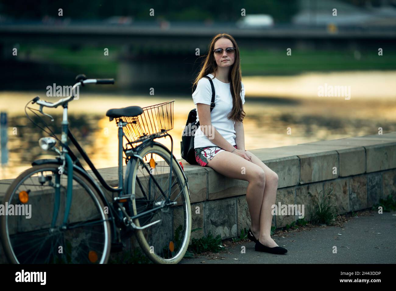 Das Teenager-Mädchen mit dem Fahrrad sitzt am Ufer des Stadtflusses. Stockfoto
