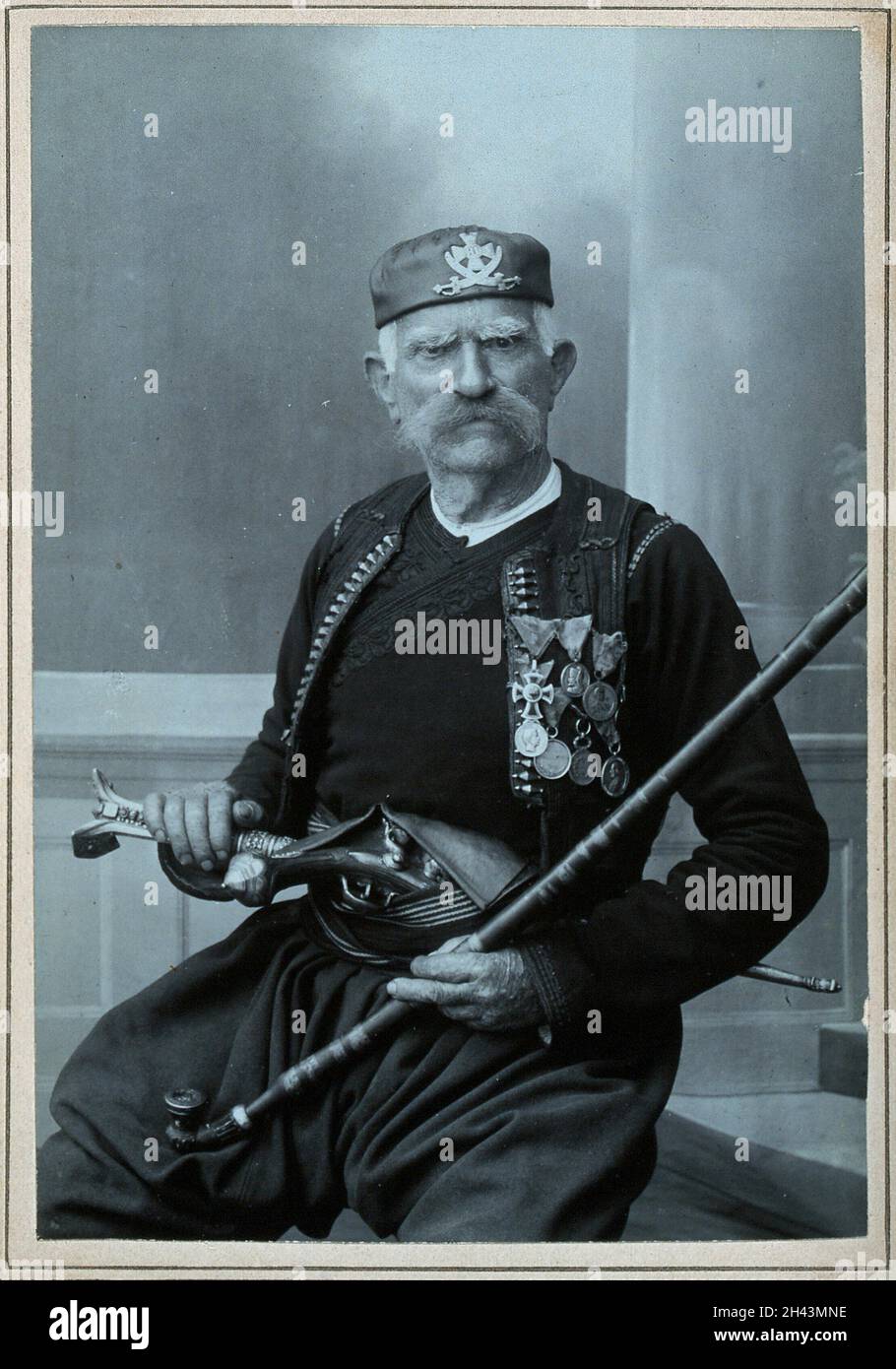 Ein älterer serbischer Mann, der in Nationaltracht mit vielen Medaillen, mit einer Pistole im Gürtel und einer Tabakpfeife in der Hand ist. Foto von D. Djordjevic, erstes Viertel des 20. Jahrhunderts. Stockfoto