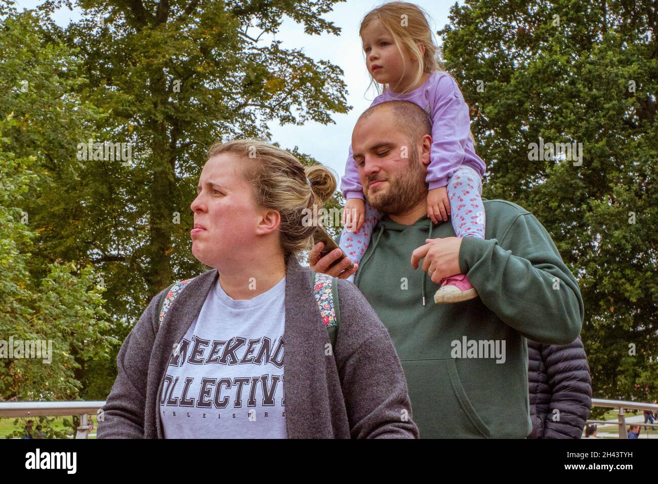 Eine junge Familie genießt einen Tag in einem Park. Vater schaut auf sein Mobiltelefon, während er seine Tochter auf seinen Schultern trägt. Stockfoto