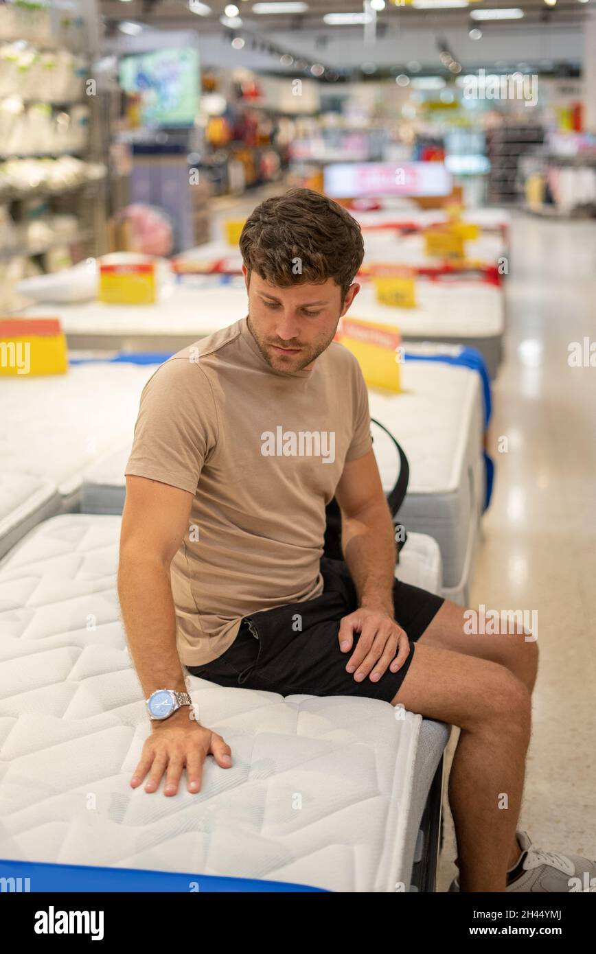 Junge Casual Kerl auf weichen Bett Matratze und berühren weiches Material sitzen, während Kommissionierung neue für zu Hause Stockfoto