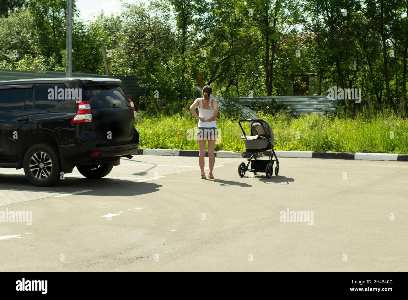 Ein Mädchen mit einem Kinderwagen auf dem Parkplatz. Eine Frau ruht in der Nähe des Autos. Eine Person nach einer langen Reise steht auf der Straße. Kinderwagen zum Spazierengehen mit einem Lauch Stockfoto