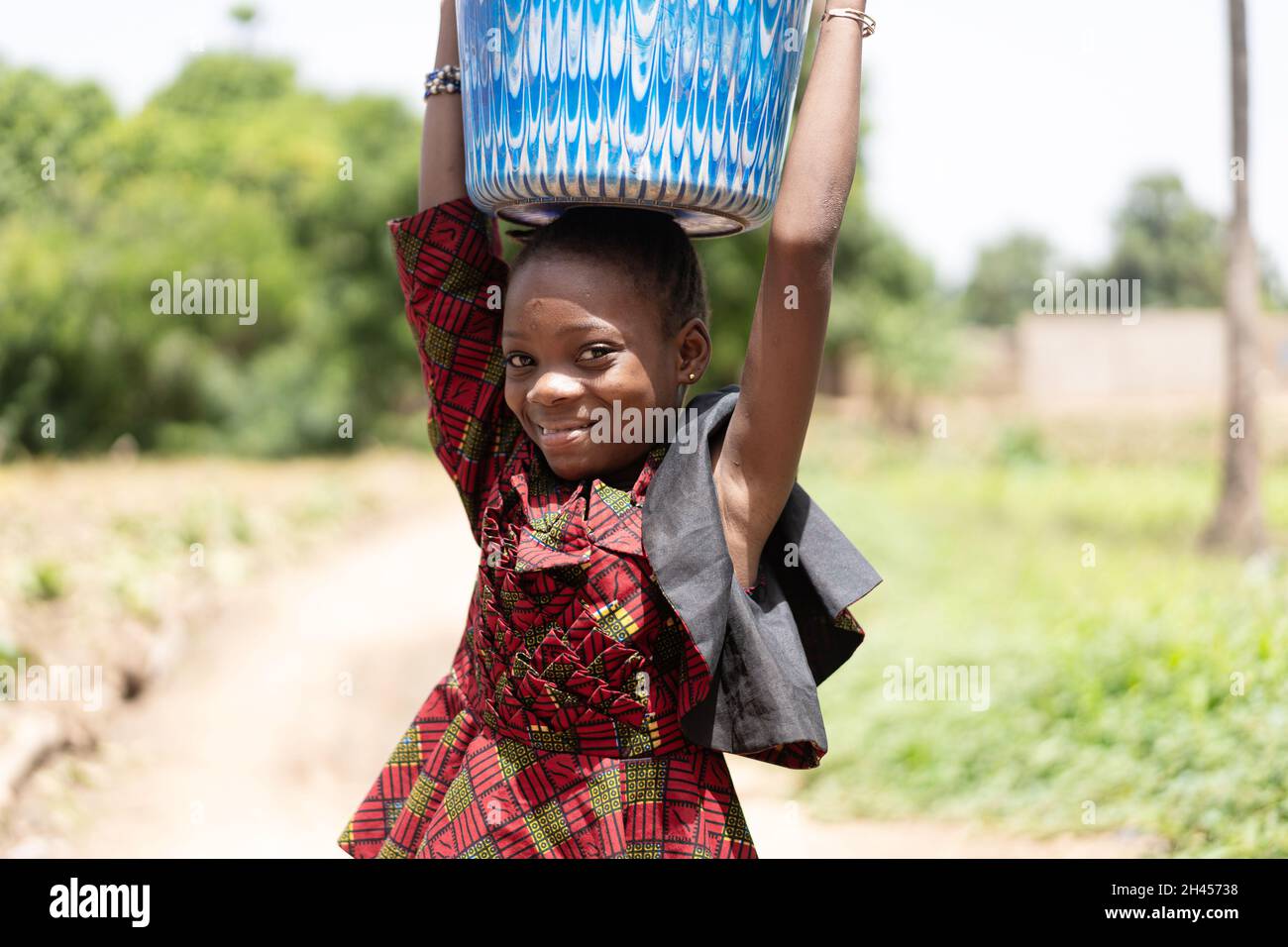 In diesem Bild balanciert ein grinsendes, georgenes schwarzes afrikanisches Mädchen einen schweren Eimer Wasser auf ihrem Kopf Stockfoto