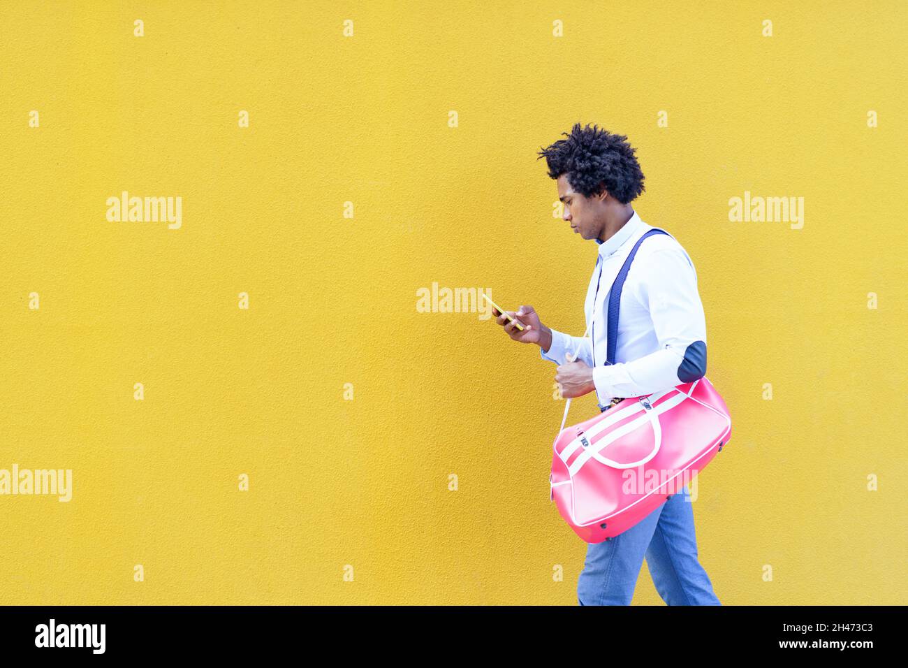Schwarzer Mann mit Afro-Frisur, der eine Sporttasche und ein Smartphone auf gelbem Hintergrund trägt. Stockfoto