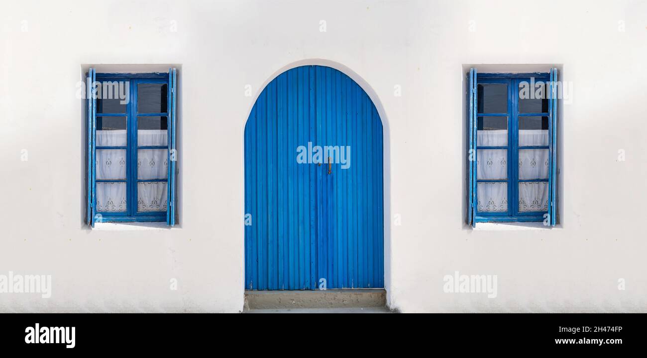 Blaue Vintage-Tür mit Bogenfenstern mit geöffneten Fensterläden und Vorhängen auf einem leeren weiß getünchten Wandhintergrund. Traditionelle griechische Insel Stockfoto