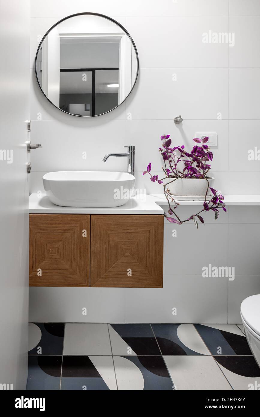 toilette, brauner Holzschrank, runder Spiegel im Waschbecken, beleuchtete Beleuchtungskörper und verschiedene Wand- und Bodenfliesen Stockfoto