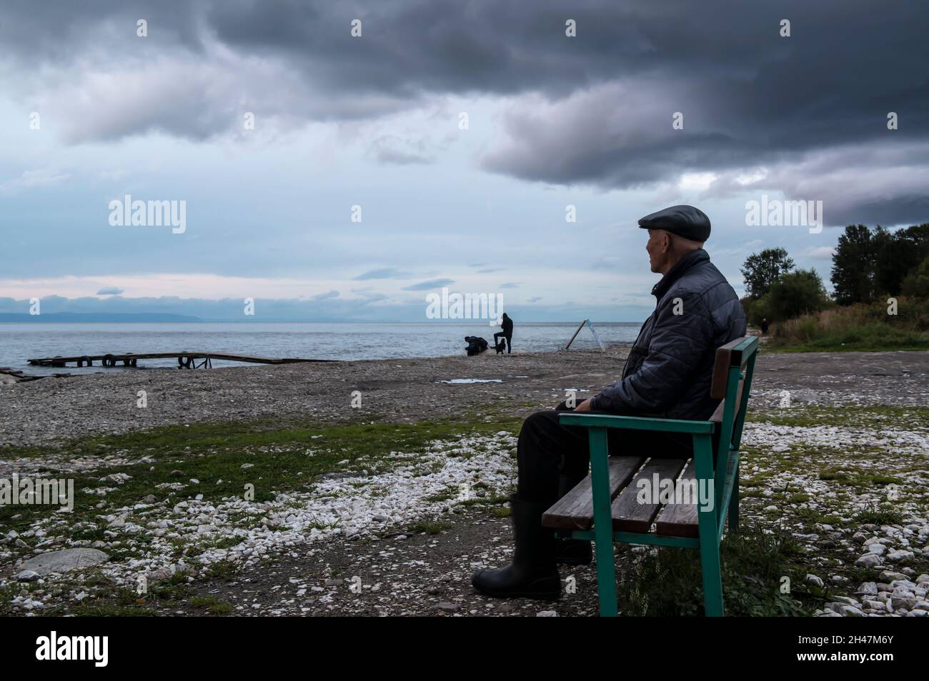 Älterer Mann, der die heranrückenden Gewitterwolken auf einer Bank am Ufer des Baikalsees beobachtet. Stadt Baikalsk, Bezirk Slyudjanski, Oblast Irkutsk, Russland Stockfoto