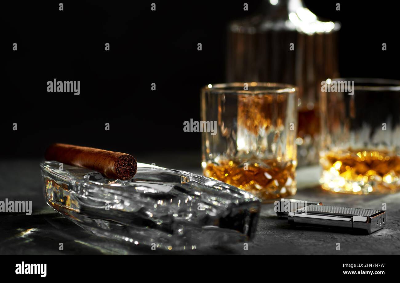 Stillleben mit einer kubanischen Zigarre in einem Aschenbecher, einem Glas und einer Flasche Whiskey und einem Feuerzeug auf einer steinernen Tischplatte. Schwarzer Hintergrund mit Platz für Text. Stockfoto