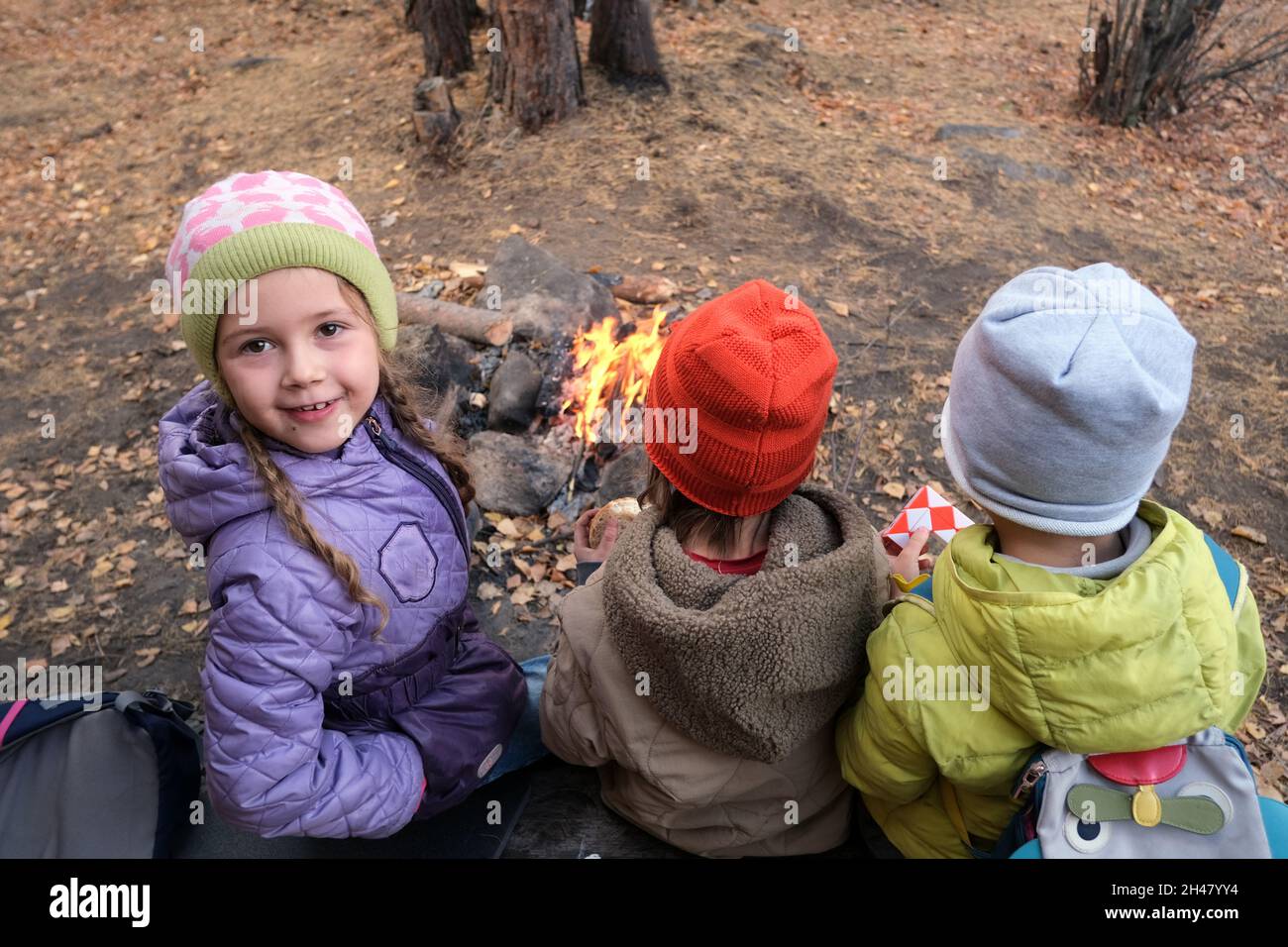 Von oben von einem positiven Mädchen in der Nähe von gesichtslosen Freunden, die während des Campens in der Nähe von brennendem Feuerholz in Herbstwäldern auf die Kamera schauen Stockfoto