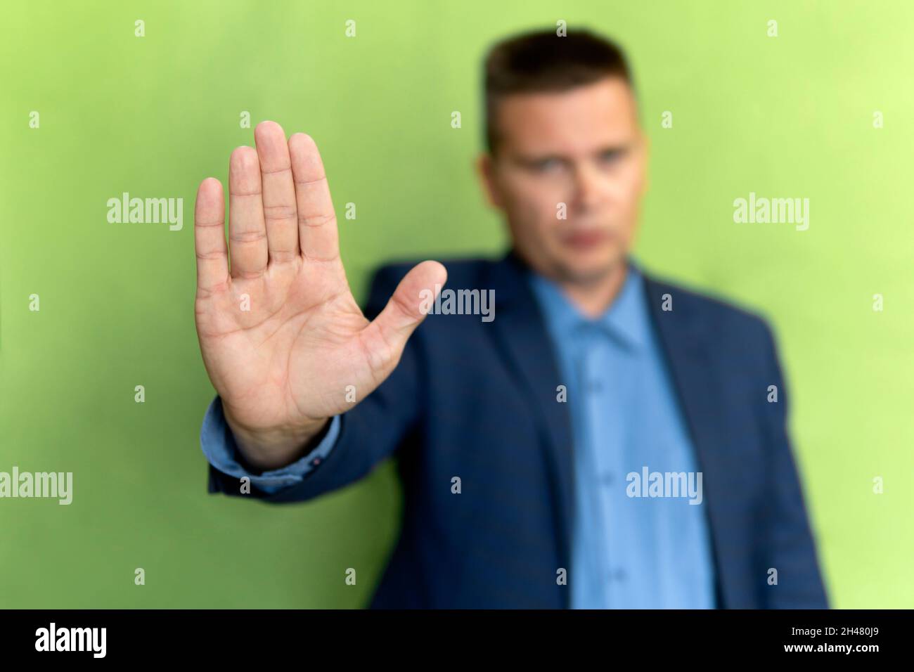 Geschäftsmann in einem blauen Anzug zeigt eine Stoppgeste auf grünem Hintergrund. Der Mann zeigt seine Handfläche. Der Wächter lässt dich nicht durch. Stockfoto