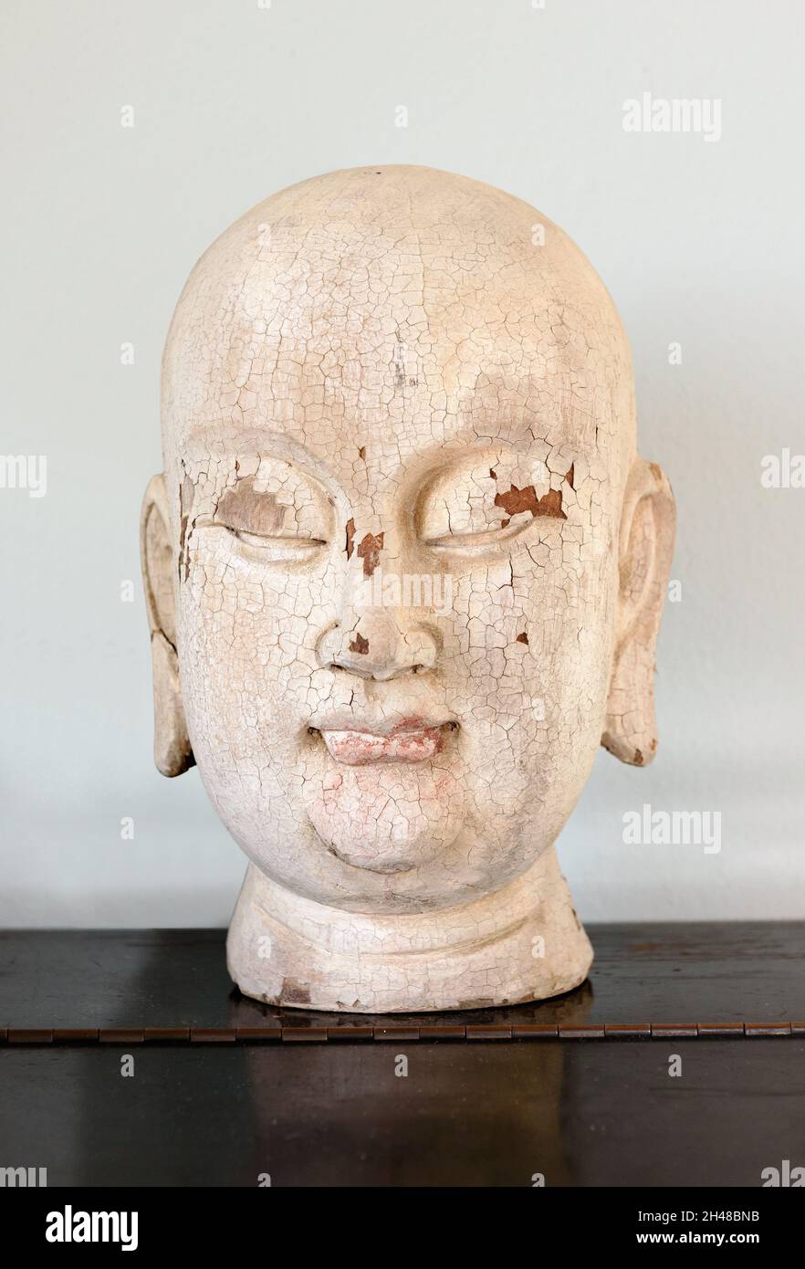 Nahaufnahme eines alten oder gealterten hölzernen Buddha-Kopfes mit knackender abblätternder Farbe und einem ruhigen spirituellen Ausdruck, der auf einem Regal dargestellt wird Stockfoto