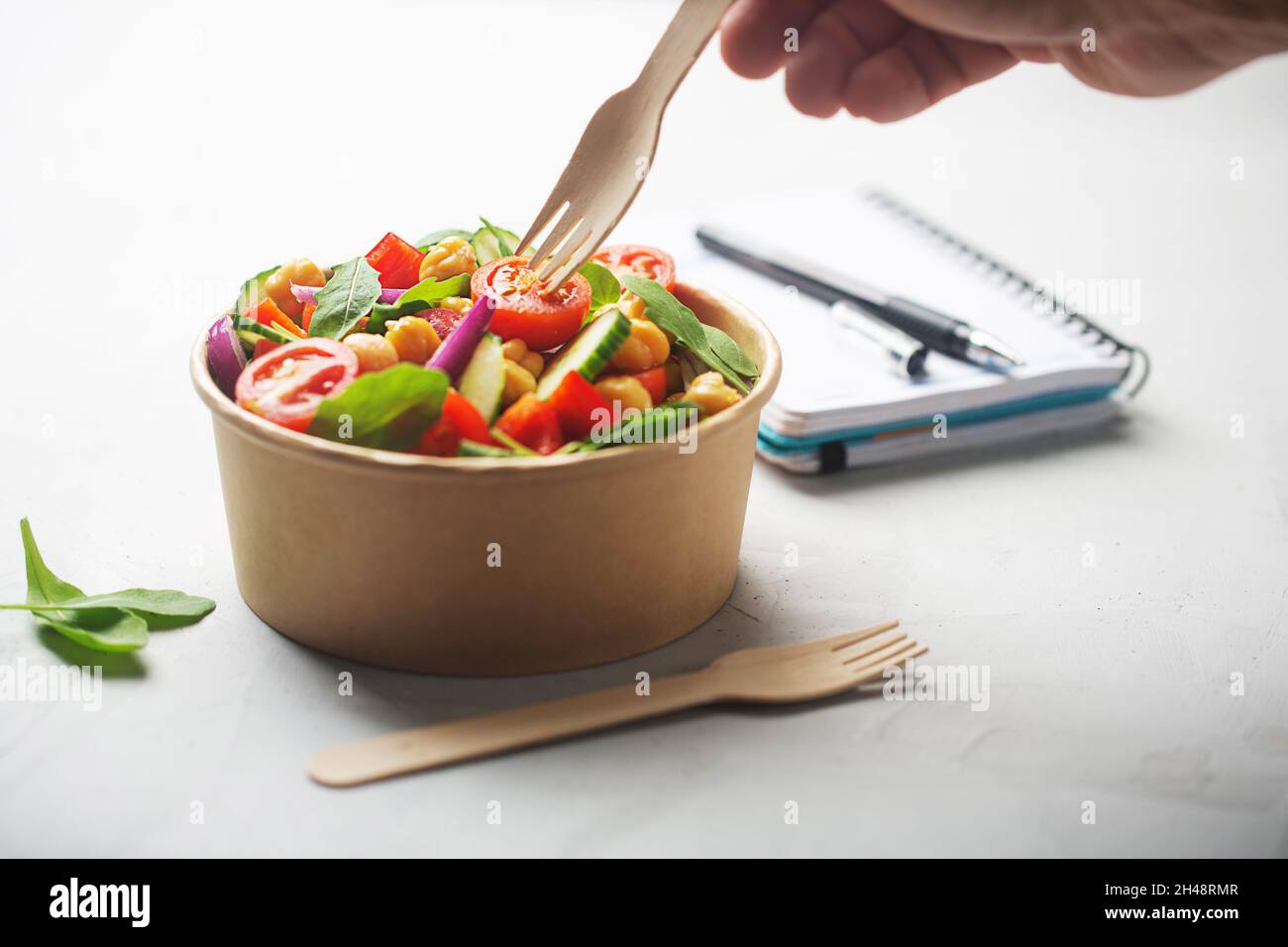 Der Mann nimmt mit einer Holzgabel Kichererbsen-Salat, der mit Tomaten, Gurken, roten Zwiebeln, Kressensalat und Rucola in einer Papierschüssel zubereitet wird. Geschirr ohne Abfall Stockfoto