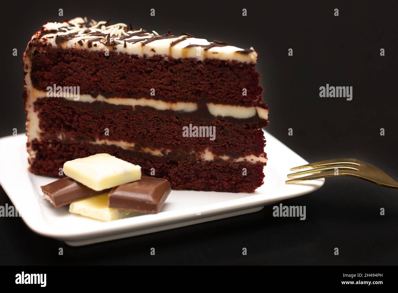 Ein Stück Schokoladenkuchen mit Creme auf einem weißen Teller, bedeckt mit Creme und weißer Schokolade auf schwarzem Hintergrund. Festliches Food-Konzept. Stockfoto