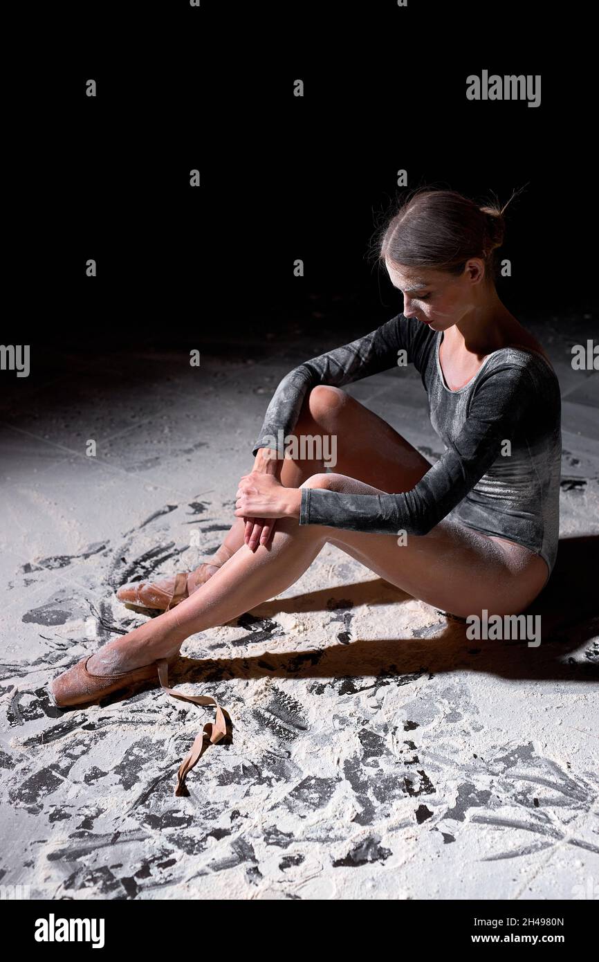 Müde junge kaukasische Frau Balletttänzerin sitzen mit Mehl auf dem Boden und machen Eine Pause allein. Liebenswert Weiblich In Schwarz Bodysuit Sitzen Isoliert Auf Bla Stockfoto