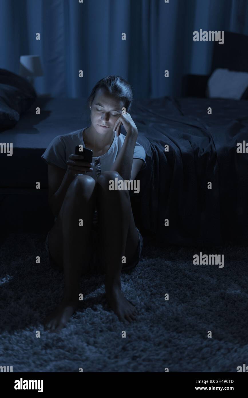 Erschöpft sitzt sie neben ihrem Bett und überprüft ihr Smartphone spät in der Nacht und leidet an Schlaflosigkeit Stockfoto