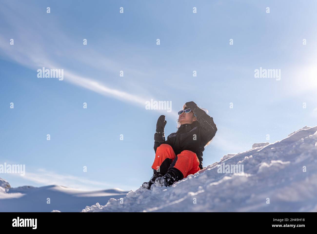 Kind sitzt im Schnee. Ein asiatisches Mädchen in Skibekleidung, Sonnenbrillen und Handschuhen im Winter. Frohe Kindheit. Stockfoto