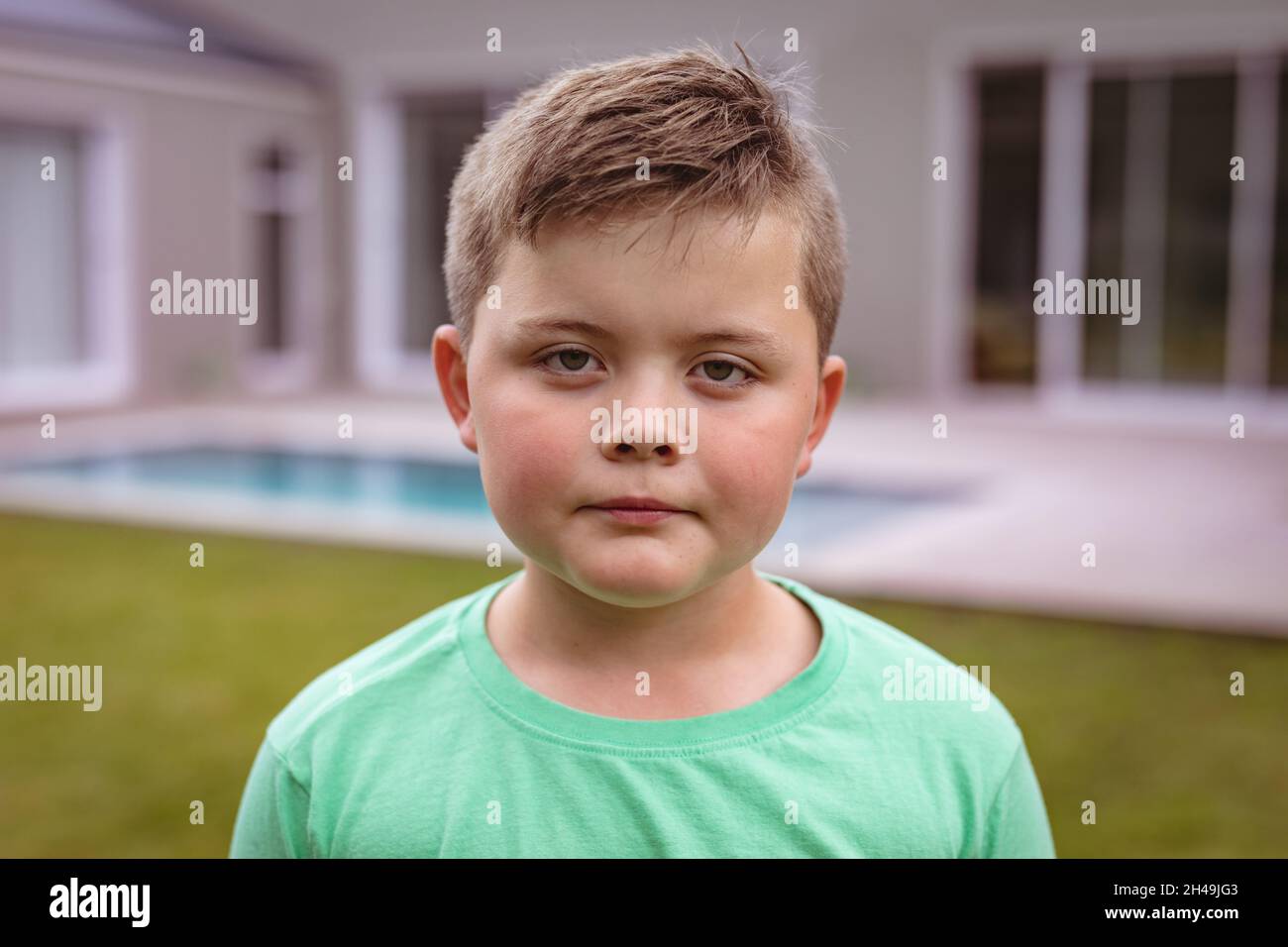 Porträt eines kaukasischen Jungen mit leerem Ausdruck, der vor dem Haus im Hinterhof steht Stockfoto