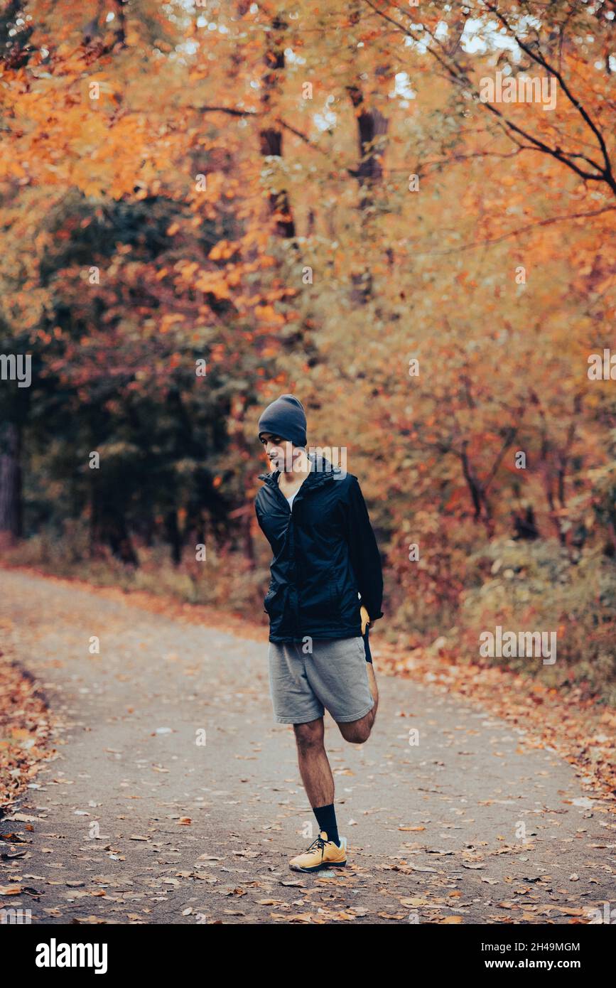 Der junge, fit schwarze Mixed-Race-Mann streckt im Herbst Quad nach oder vor einem Lauf im Wald auf einem gepflasterten Trail. Aktive Kleidung tragen. Gesunder Mann beim Laufen Stockfoto