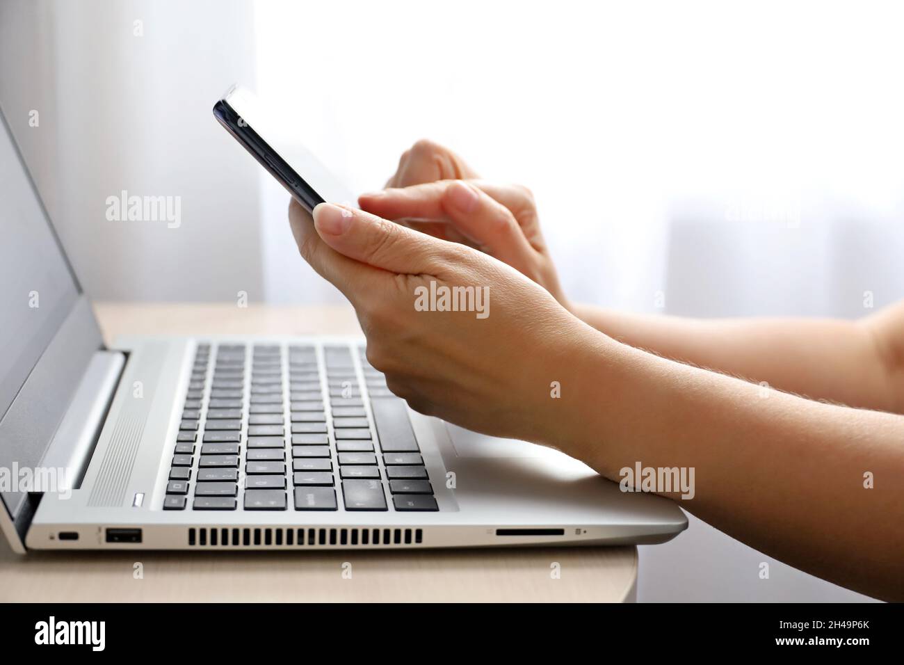 Frau mit Smartphone sitzt auf Laptop-Tastatur in der Nähe des Fensters. Konzept der Online-Kommunikation, Büro- oder Heimarbeit und Bezahlung Stockfoto