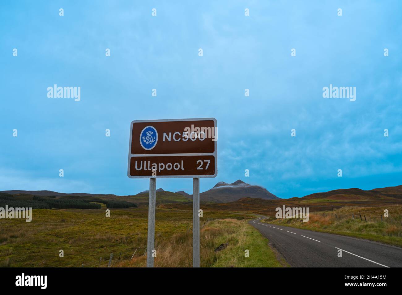 NC500 Schild mit Kilometerstand nach Ullapool. Weg zur Seite und verschwommene Berglandschaft im Hintergrund. Blauer Himmel mit Wolken, viel Kopierplatz. Stockfoto