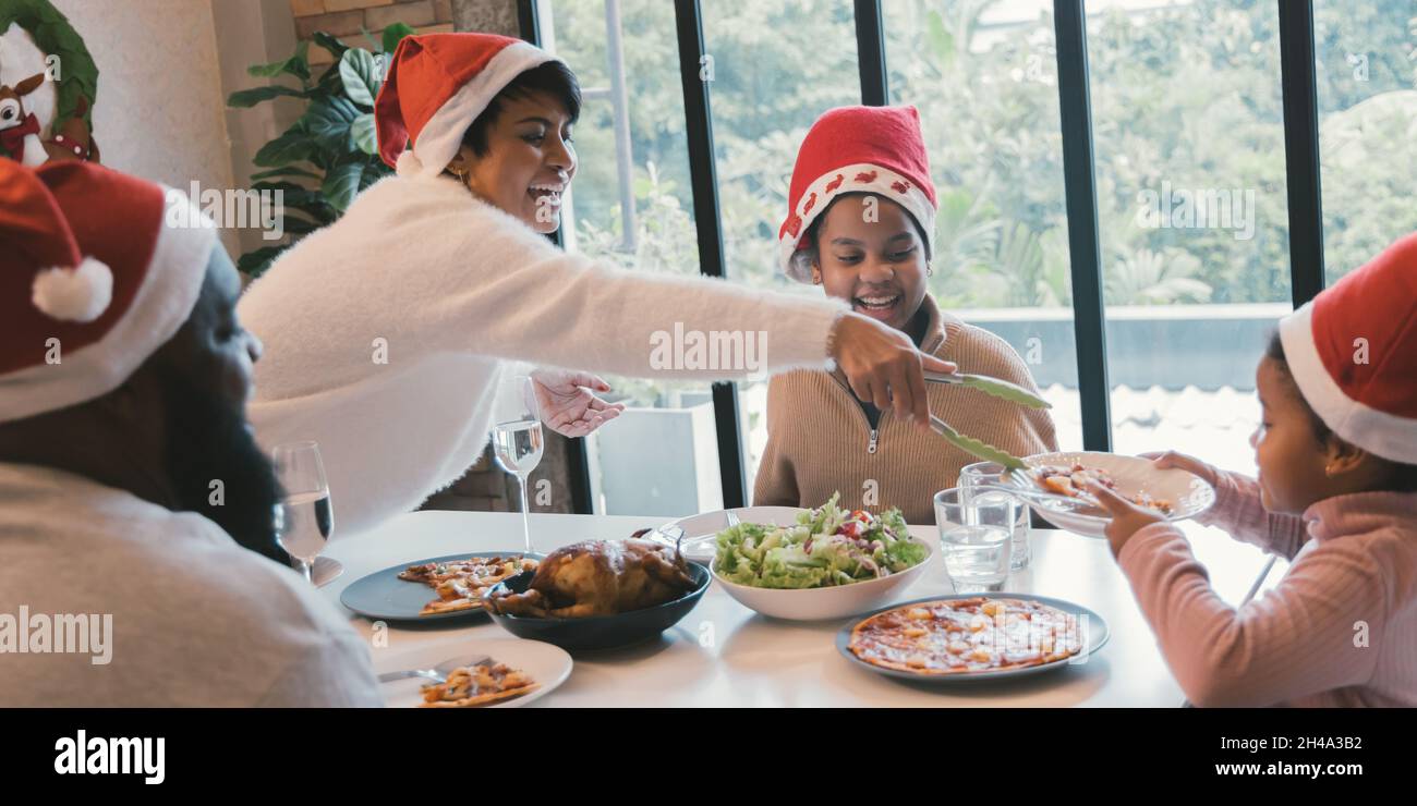 Schöne junge afroamerikanische Familie mit Mutter, Vater und zwei Töchtern, die Weihnachtsmütze tragen und das weihnachtsessen mit Pizza und truthahn zu Hause genießen Stockfoto
