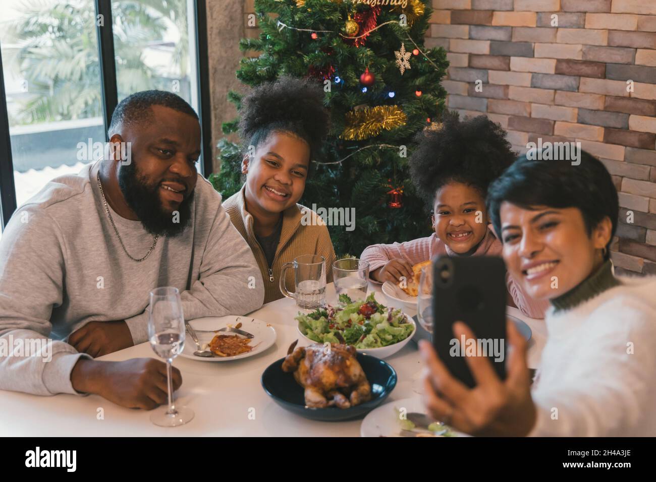 Fröhliche afroamerikanische Familie genießt weihnachten und das Mittagessen im neuen Jahr, während die Frau Selfie auf dem Smartphone mit Familienmitgliedern zu Hause gegen geschmückten Weihnachtsbaum nimmt Stockfoto