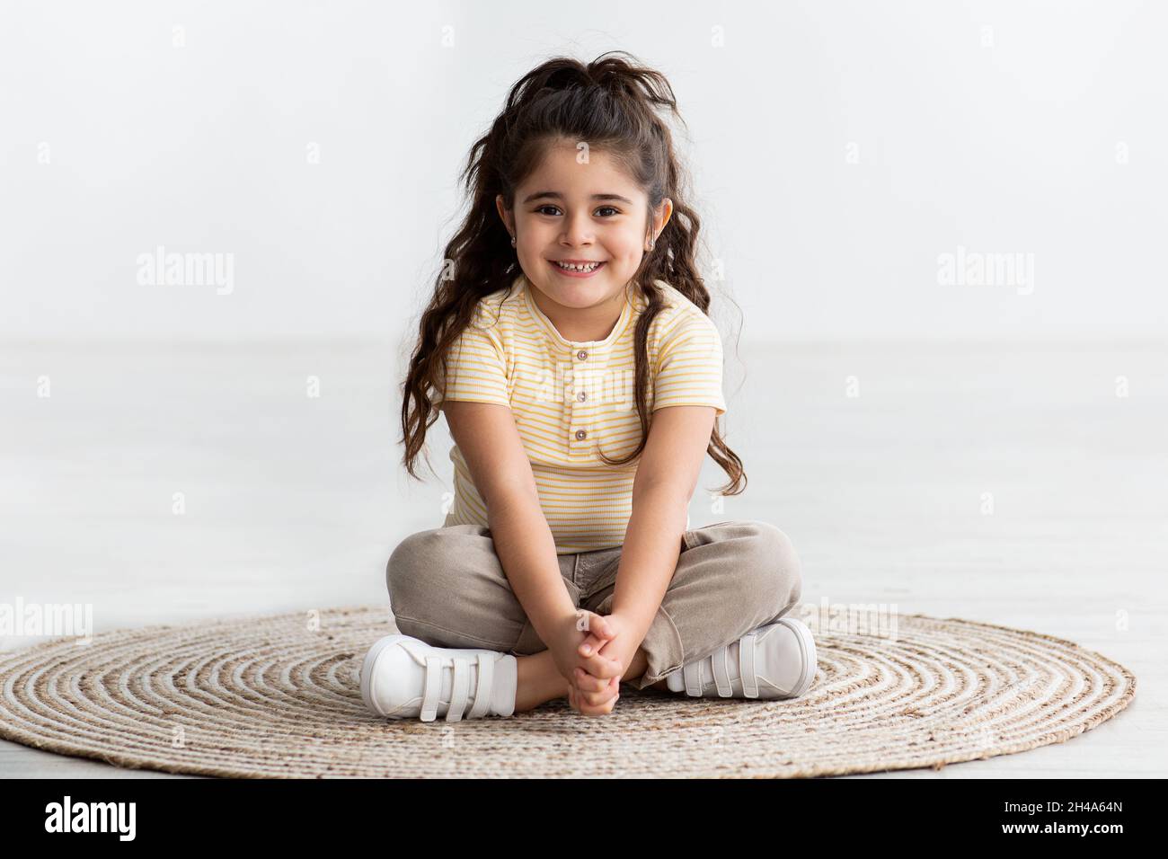 Portrait of Cute Smiling Little Girl sitzend mit Beinen gekreuzt auf Wicker Teppich zu Hause, schöne fröhlich entzückende weibliche Kind entspannend auf dem Boden, L Stockfoto