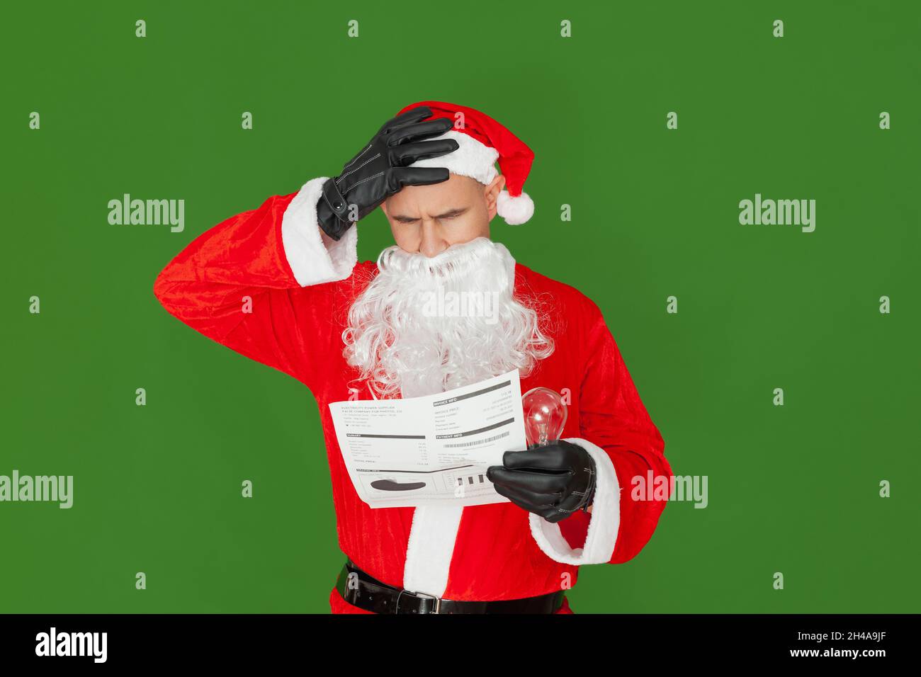 Ein Mann, der als Weihnachtsmann gekleidet ist, hält einen Stromschein und eine Glühbirne in der einen Hand, während er die andere Hand besorgt an seinen Kopf hält. Der Hintergrund i Stockfoto