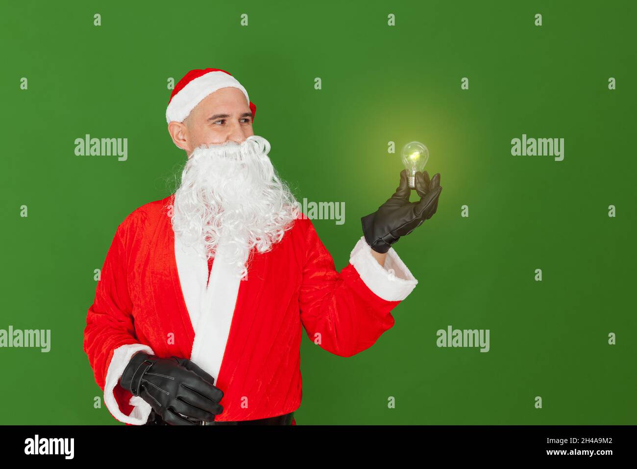 Ein Mann, der als Weihnachtsmann gekleidet ist, hält eine Glühbirne mit einer Hand, die leuchtet und Licht ausstrahlt. Der Hintergrund ist grün. Stockfoto
