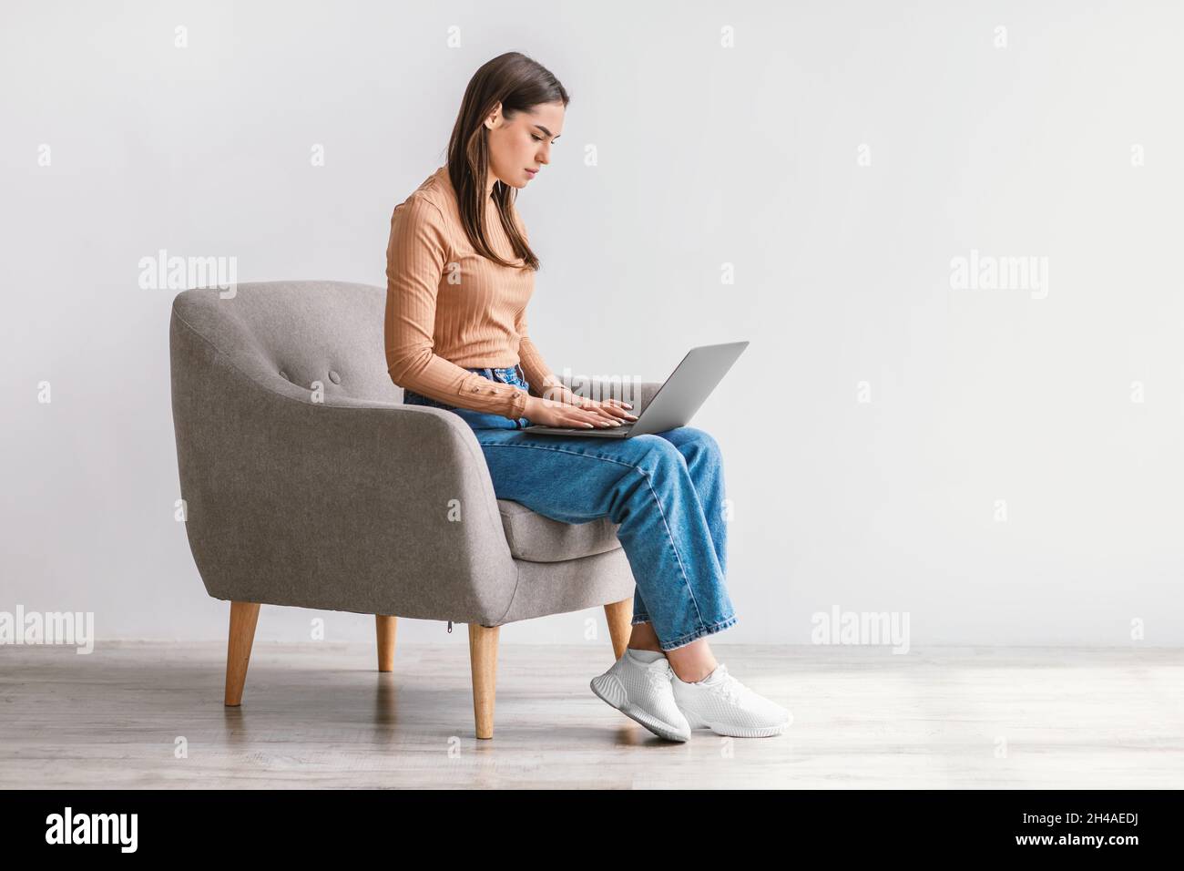 Seriöse Millennial-Dame, die im Sessel mit Laptop sitzt, online studiert oder Videokonferenz gegen weiße Studiowand, leerer Raum. Junge Cauc Stockfoto