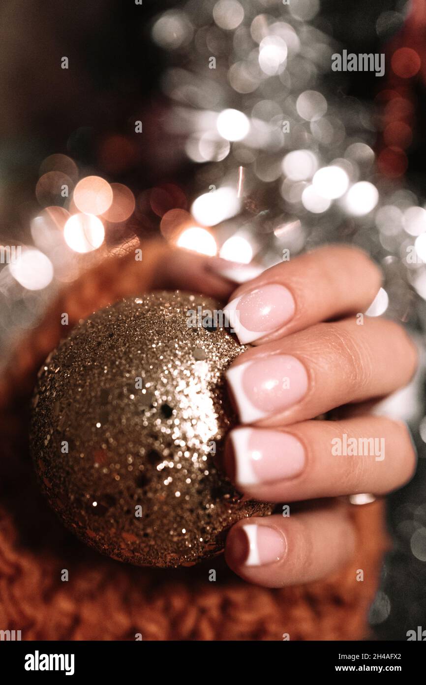 Weibliche Hand mit französischer Maniküre, die einen goldenen Weihnachtsball hält. Winterfeiertag. Neujahrskonzept. Stockfoto