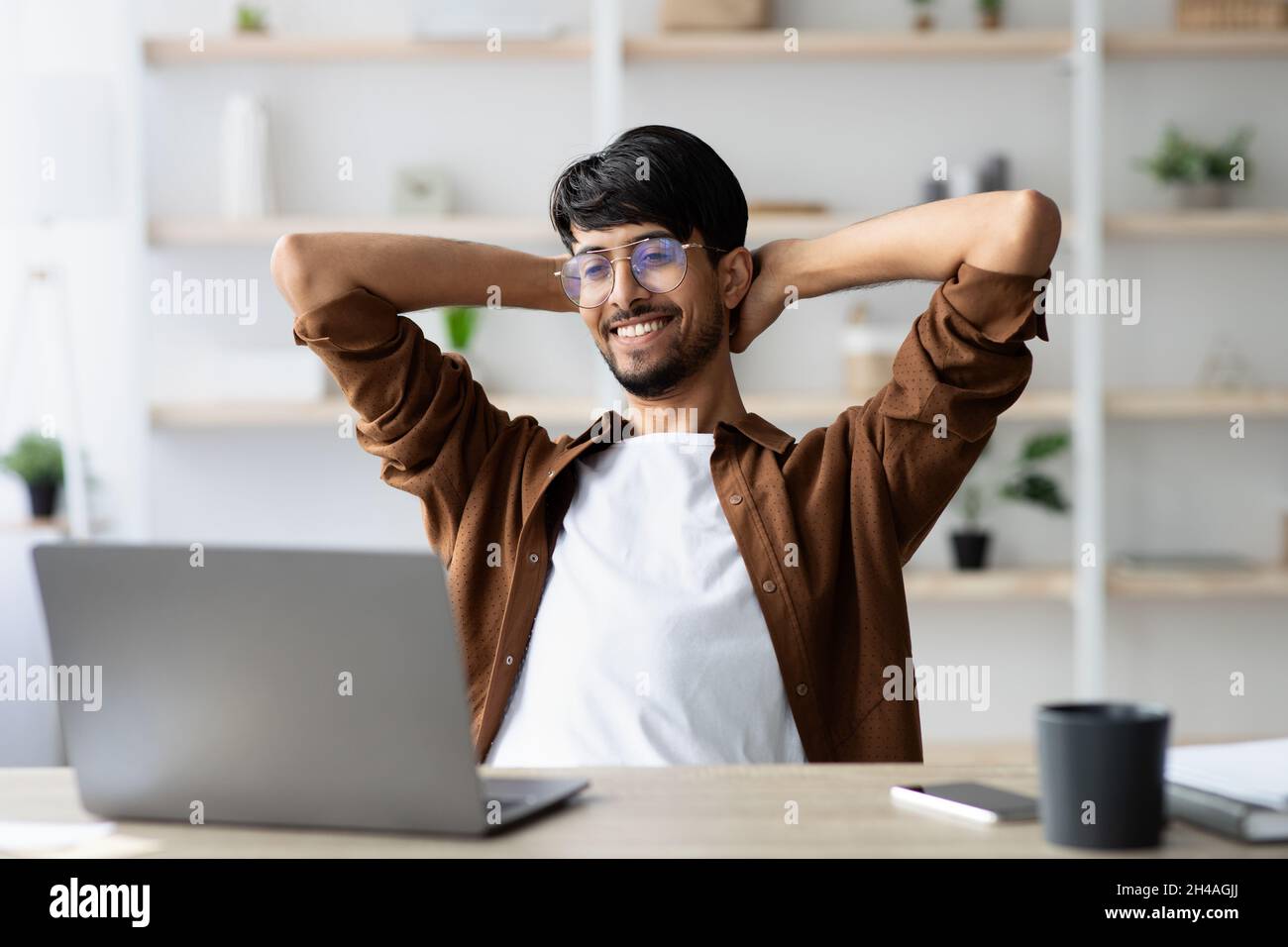 Wohlhabender indischer Projektmanager, der vor einem modernen Laptop am Arbeitstisch sitzt, sich zurücklehnt und lächelt, sich gut fühlt, was seinen Job anstellt, Bürointeri Stockfoto