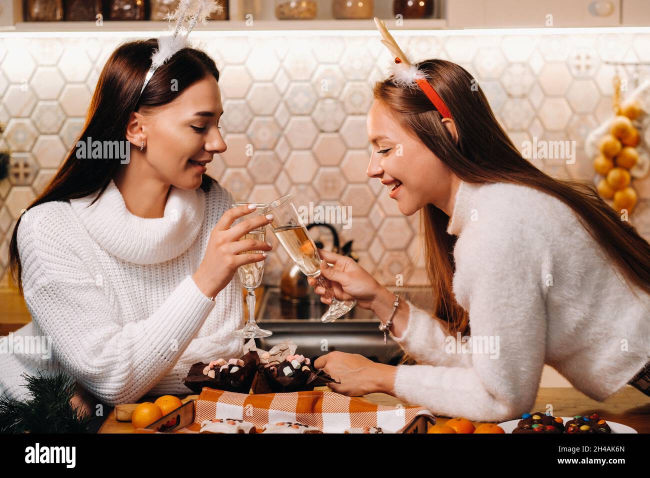 Zwei Mädchen in einer gemütlichen häuslichen Umgebung mit Champagner in den Händen zu Weihnachten. Lächelnde Mädchen trinken Champagner an einem festlichen Abend. Stockfoto