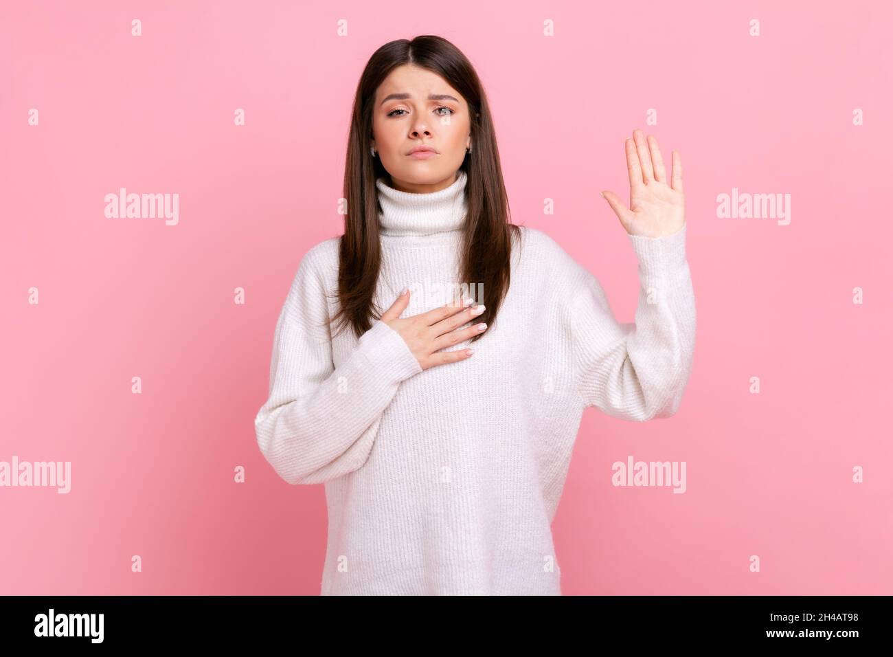 Porträt einer ehrlichen jungen Frau, die mit der Hand auf der Brust und den Fingern nach oben steht und Loyalität verspricht, in einem weißen Pullover im lässigen Stil. Innenaufnahme des Studios isoliert auf rosa Hintergrund. Stockfoto