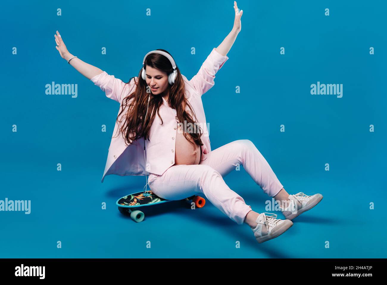 Eine Schwangere im pinken Anzug sitzt mit Kopfhörern auf einem Skateboard und hört Musik auf blauem Hintergrund. Stockfoto