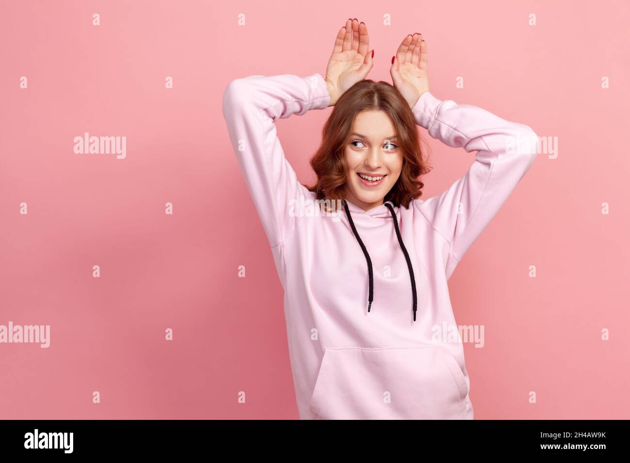 Porträt einer unbeschwerten glücklichen Brünette junge Frau im Kapuzenpullover, die lustige Hasenohren mit Händen am Kopf macht, kindhaftes Verhalten, verspielte optimistische Stimmung. Innenaufnahme des Studios isoliert auf rosa Hintergrund Stockfoto