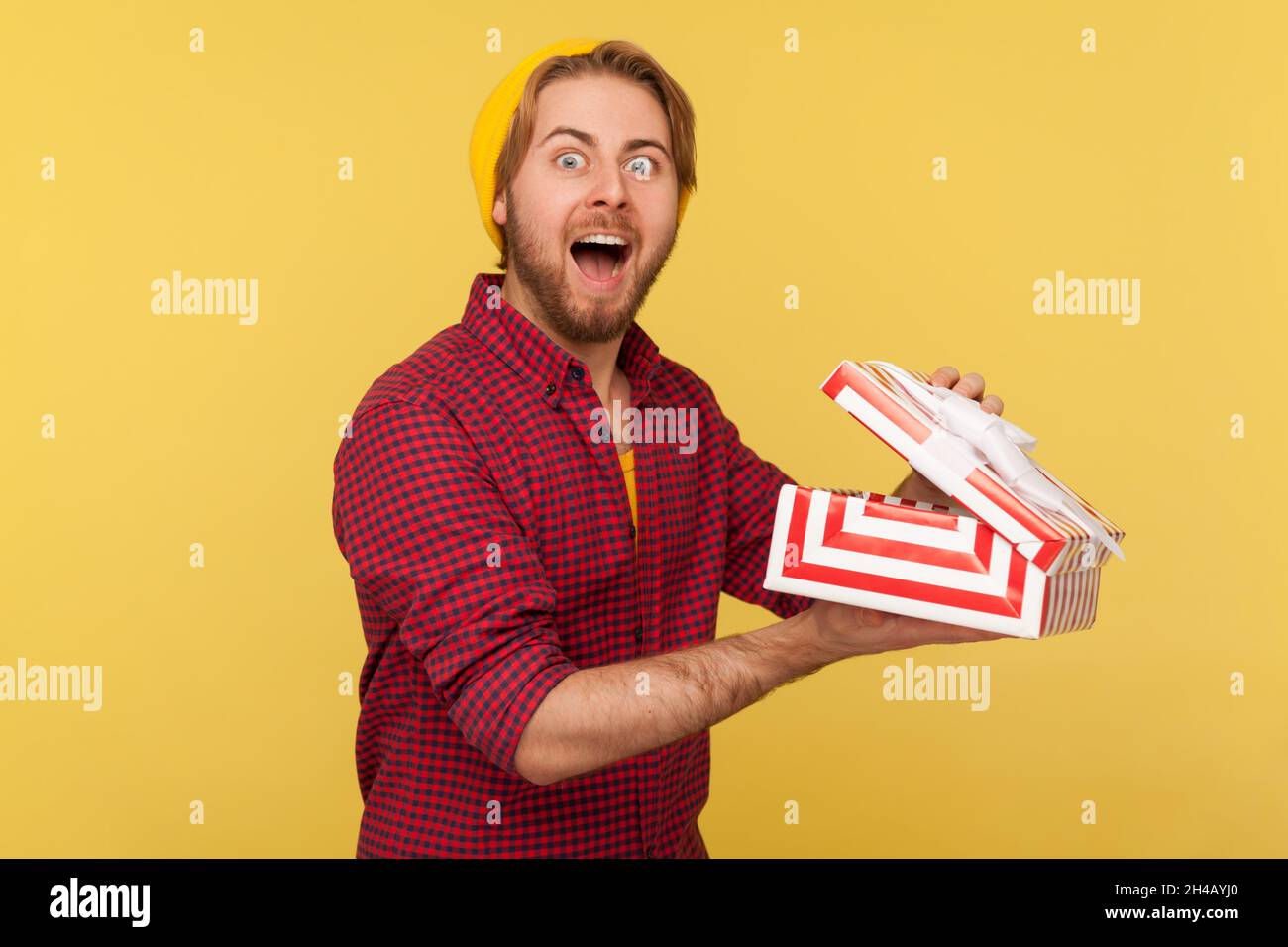 Porträt eines bärtigen Mannes mit Hipster in Beanie-Hut und kariertem Hemd, der eine geöffnete Geschenkbox hält und mit einem erstaunten Lächeln auf die Kamera blickt und dabei das Geschenk auspacken kann. Innenaufnahme des Studios isoliert auf gelbem Hintergrund. Stockfoto