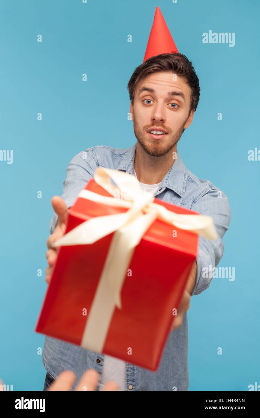 POV Hand geben verpackt rote Geschenkbox zu bärtigen Mann mit Party-Kegel auf dem Kopf, Kerl trägt Denim-Shirt Blick auf Geburtstagsgeschenk Überraschung mit Freude. Innenaufnahme des Studios isoliert auf blauem Hintergrund. Stockfoto