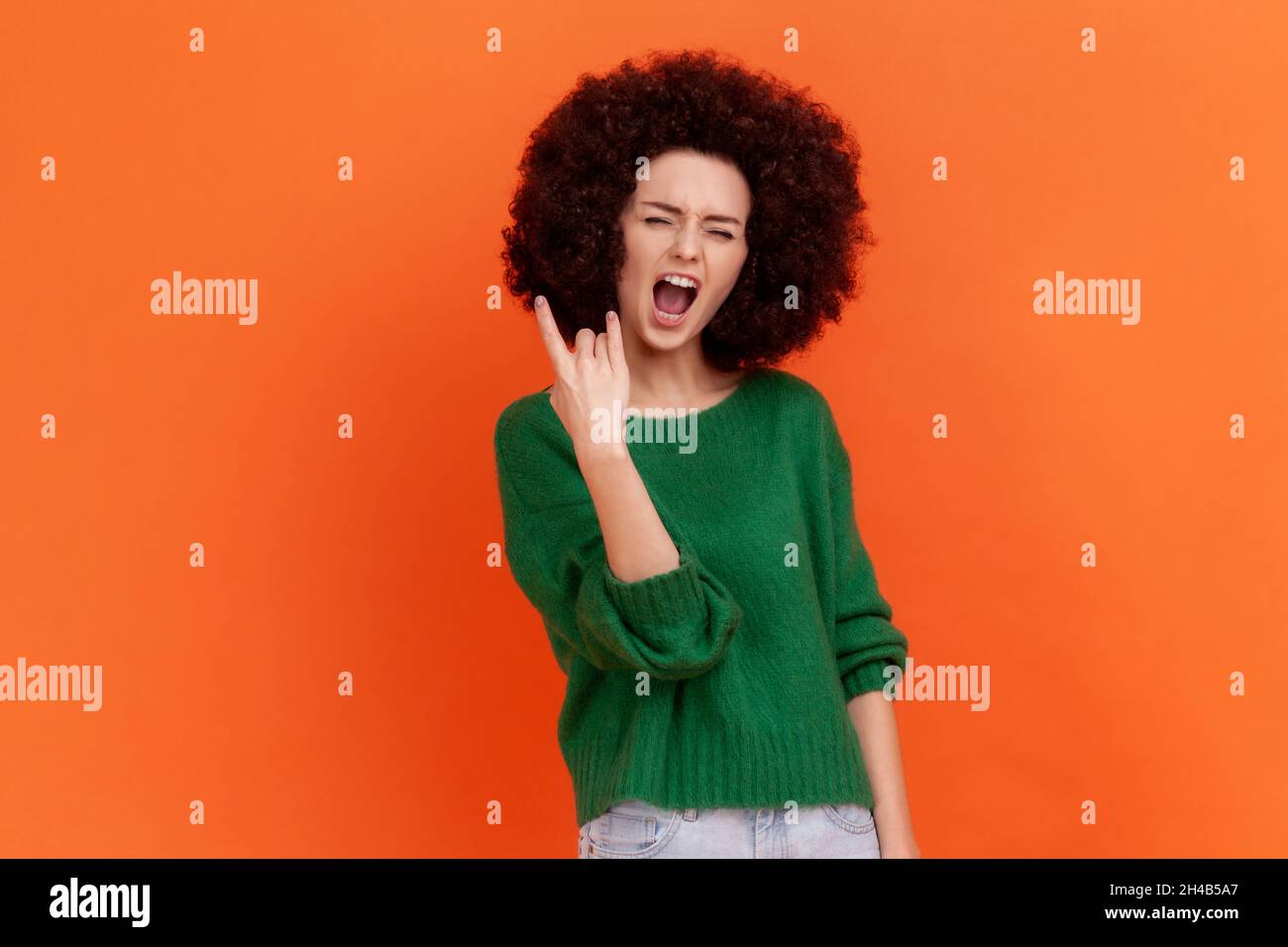 Aufgeregt enthusiastische Frau mit Afro-Frisur trägt grünen lässigen Stil Pullover mit Rock and Roll Geste. Schreiend begeistert, Indoor Studio Schuss isoliert auf orangefarbenem Hintergrund. Stockfoto