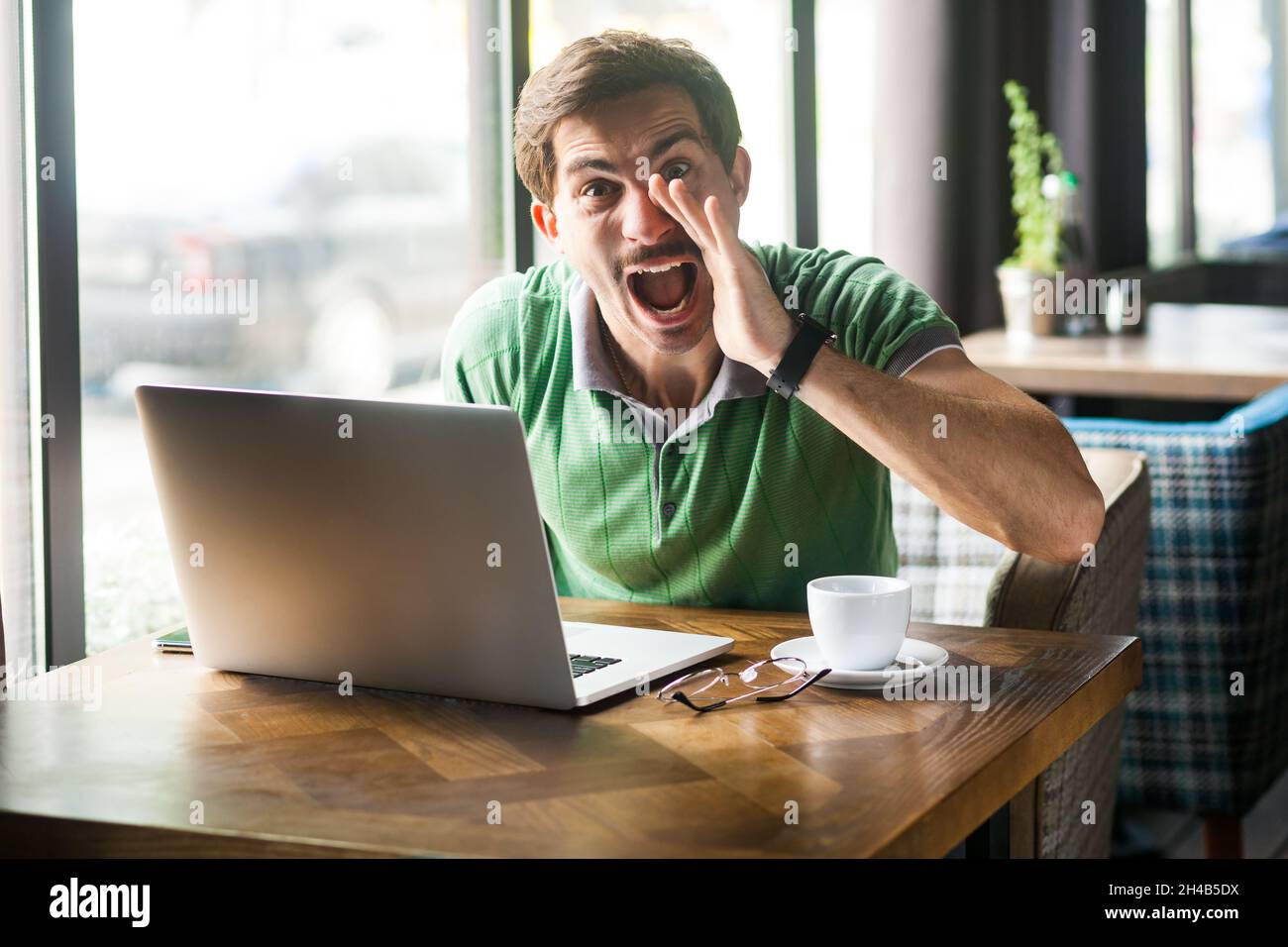 Porträt eines verrückten Mannes mit grünem T-Shirt, sitzend mit der Hand neben dem Mund und schreiend, um etwas zu teilen, während er online am Laptop arbeitet, Innenaufnahme in der Nähe eines großen Fensters, Café-Hintergrund. Stockfoto