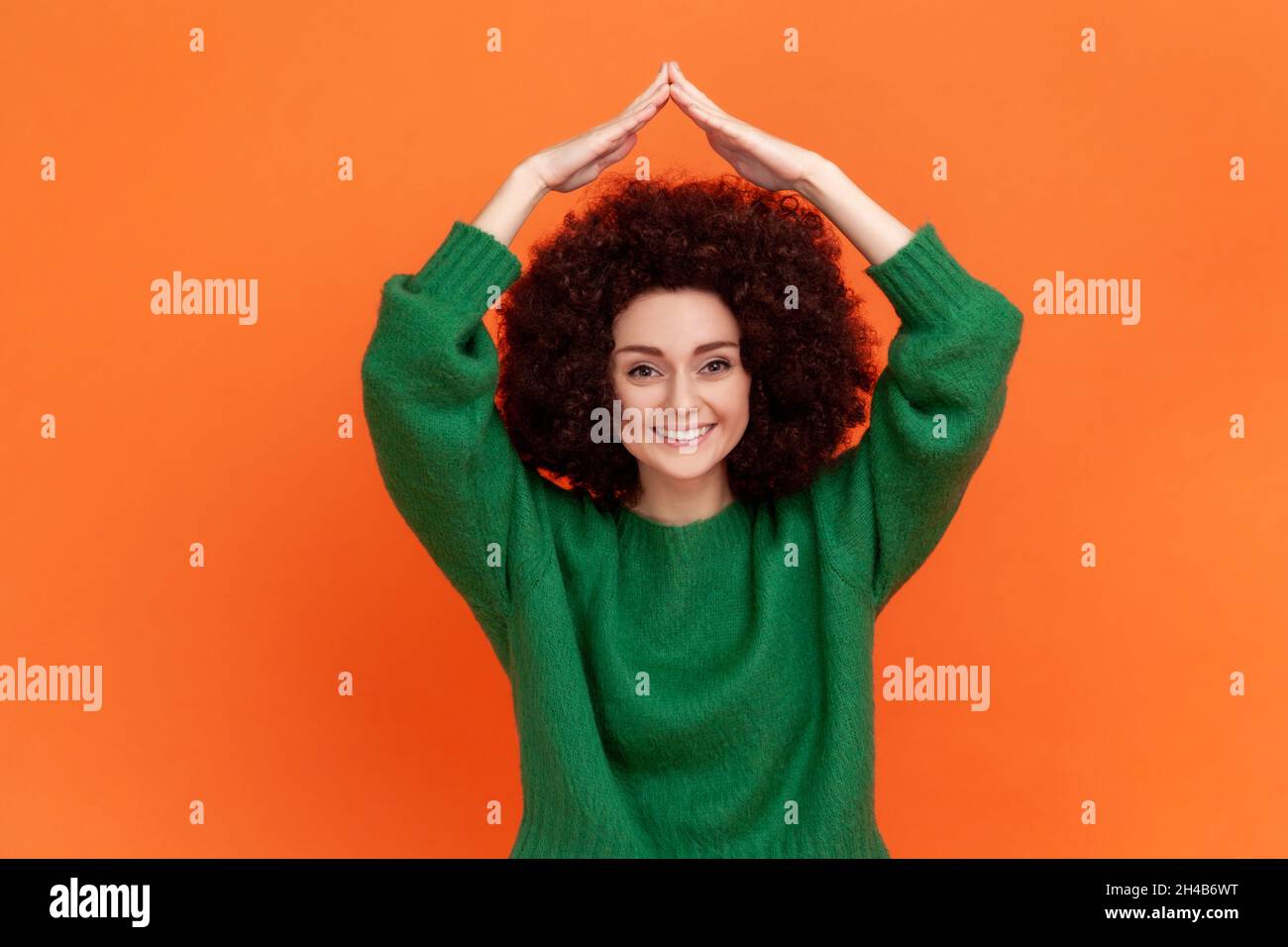 Frau mit Afro-Frisur trägt grünen lässigen Stil Pullover machen Dach Geste und lächeln, Konzept der Lebensversicherung, Schutz. Innenaufnahme des Studios isoliert auf orangefarbenem Hintergrund. Stockfoto