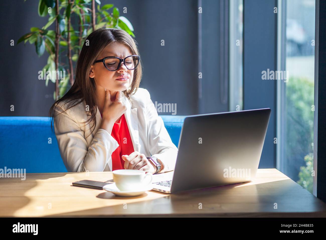 Porträt einer ungesunden Frau in einer Brille, die am Laptop arbeitet, schmerzhaften Hals hält, Halsschmerzen, entzündete Mandeln, Grippesymptome leidet. Innenaufnahme, Café oder Bürohintergrund. Stockfoto