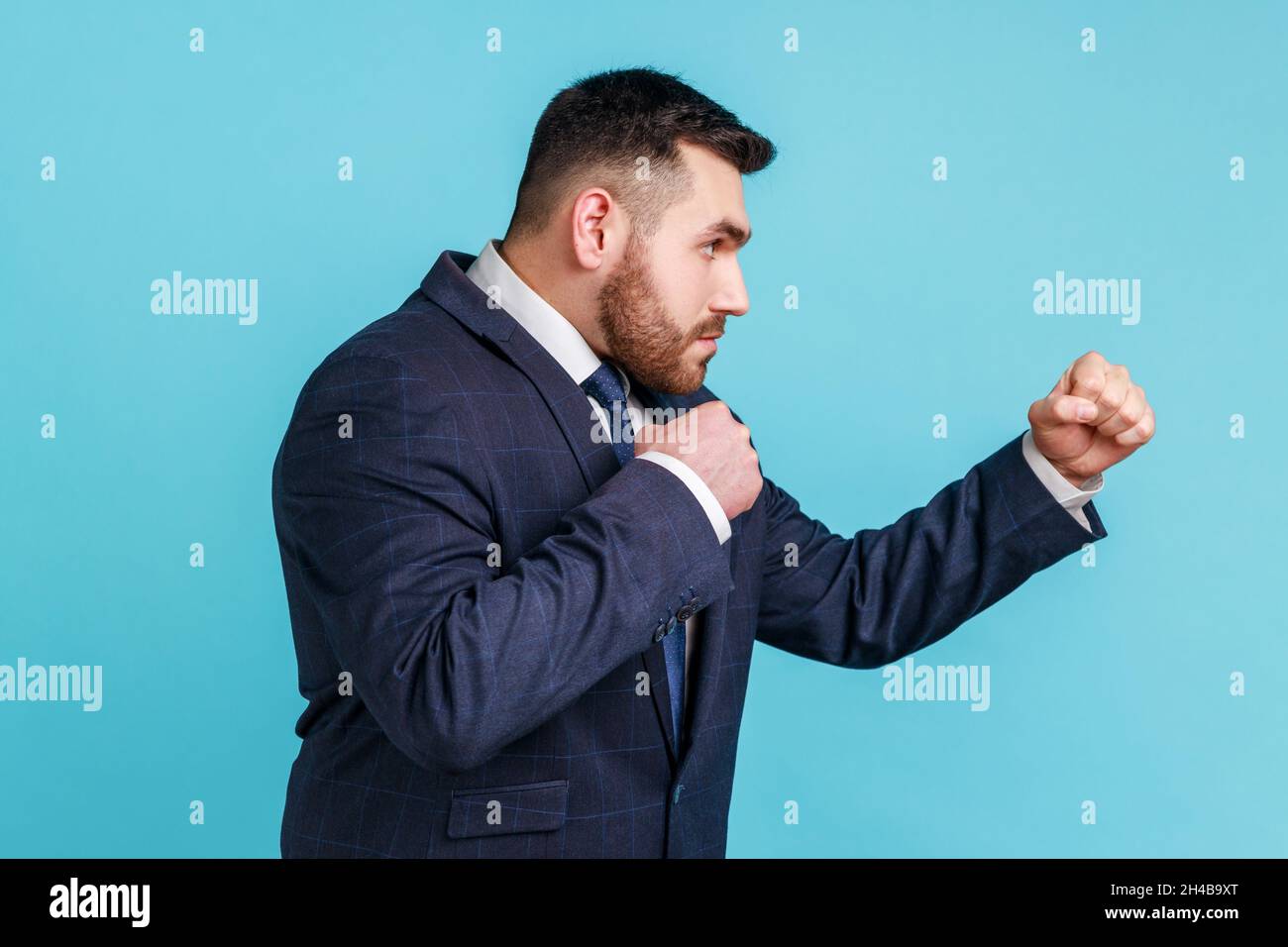 Seitenansicht eines aggressiven Geschäftsmannes, der im offiziellen Stil einen Anzug trägt, der zum Stanzen bereit ist, mit geballten Fäusten boxt und mit jemandem kämpft. Innenaufnahme des Studios isoliert auf blauem Hintergrund. Stockfoto