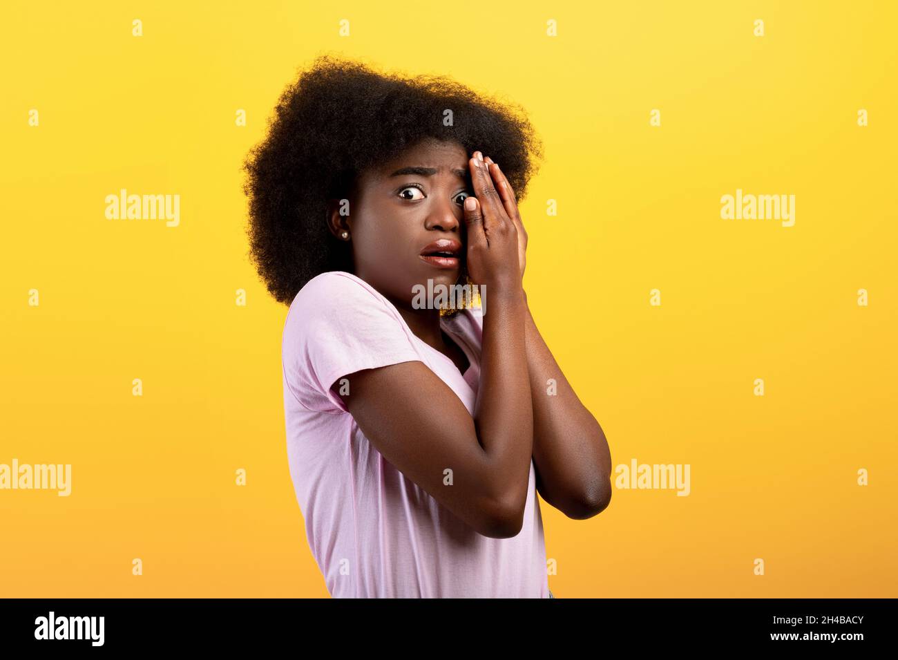 Gestresste junge schwarze Dame, die das Gesicht mit Händen bedeckt und mit einem Auge guckt, sich ängstlich fühlt und auf gelbem Studiohintergrund steht. Verängstigte Frau tr Stockfoto
