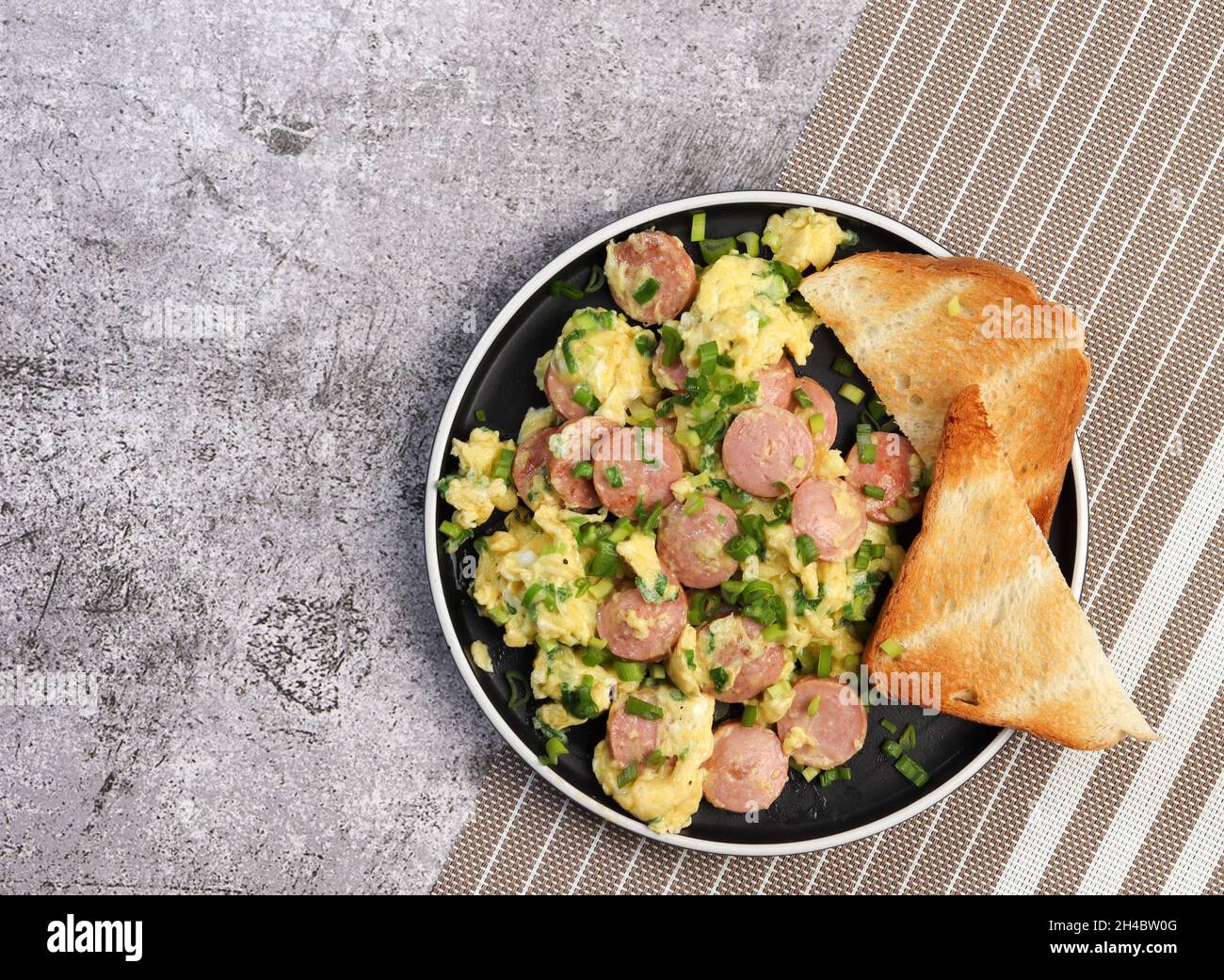 Rührei mit Wurst und Toast auf einem runden Teller auf dunklem Grund. Draufsicht, flach liegend Stockfoto