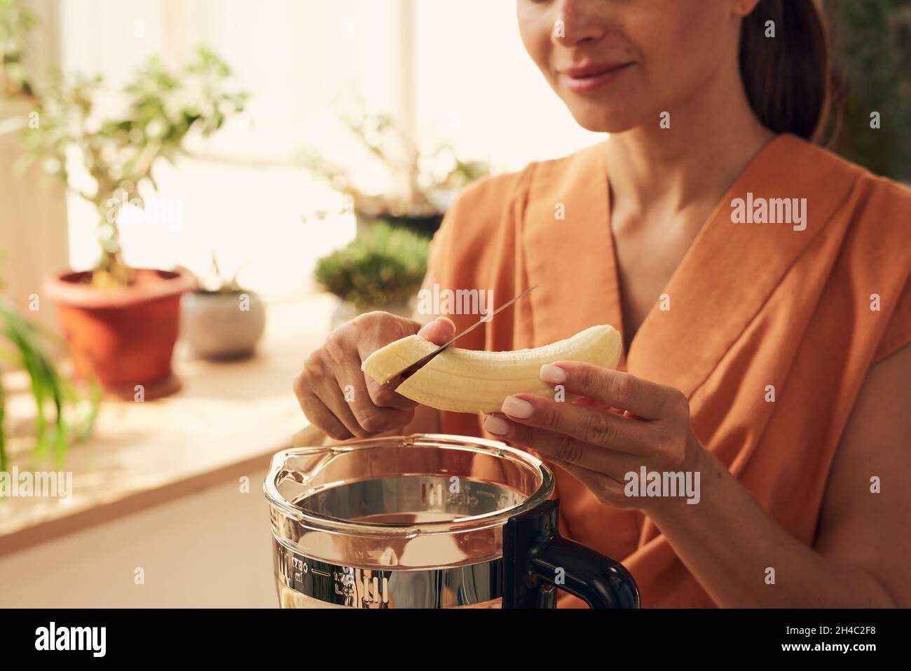 Junge Frau hackt geschälte Banane über den elektrischen Mixer, während sie hausgemachten Smoothie für das Familienfrühstück zubereitet Stockfoto